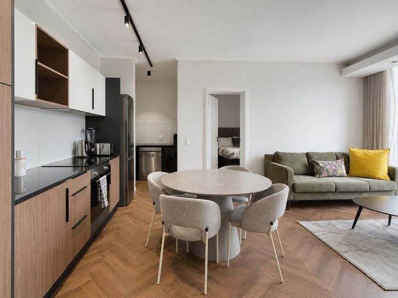 Modern open-plan living area featuring a sleek kitchen with wood-tone cabinetry, stone countertops, and herringbone timber flooring, seamlessly flowing into the dining and lounge space.