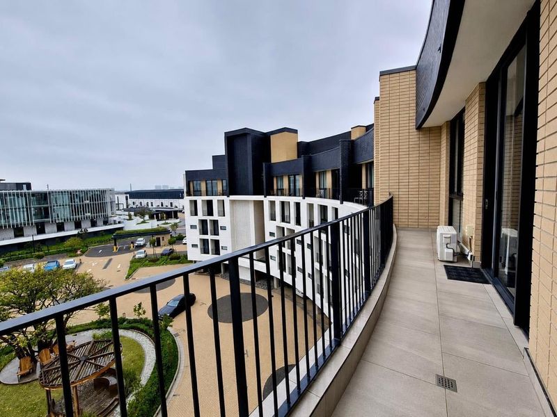 A spacious curved balcony with tiled flooring, metal railings, and views overlooking the apartment complex courtyard and surrounding development.