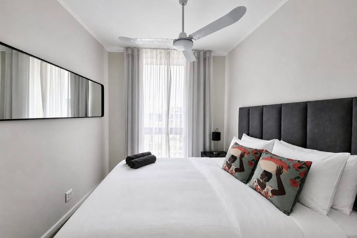 A well-maintained bedroom featuring a ceiling fan, neutral wall finishes, and large windows with sheer curtains, providing a bright and comfortable living space.