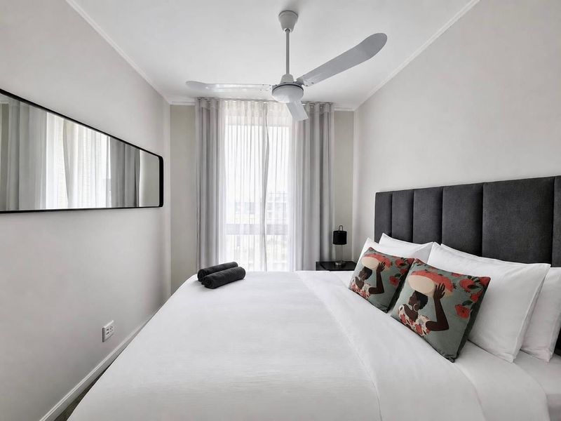 A well-maintained bedroom featuring a ceiling fan, neutral wall finishes, and large windows with sheer curtains, providing a bright and comfortable living space.