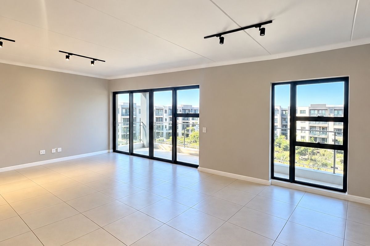A bright, spacious, and empty living area featuring neutral tiled floors, modern track lighting, and large windows with access to a balcony.