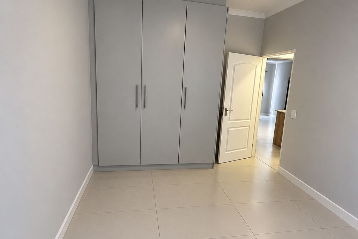 A bright, empty bedroom featuring neutral tiled flooring, modern grey built-in wardrobes, and clean, freshly painted walls.