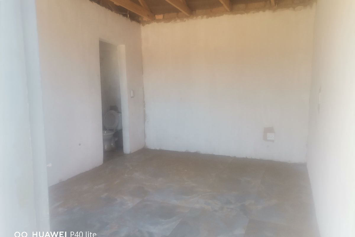 An unfinished bedroom space featuring exposed timber roof trusses, tiled flooring, and an adjacent en-suite bathroom area.