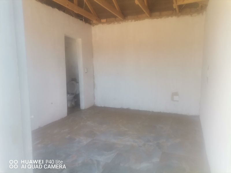 An unfinished bedroom space featuring exposed timber roof trusses, tiled flooring, and an adjacent en-suite bathroom area.
