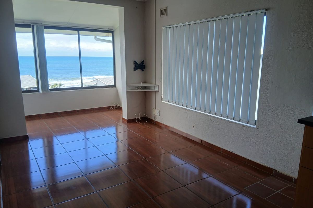 An empty living room featuring tiled flooring, vertical blinds, and a wall-mounted TV bracket, with large windows offering a scenic ocean view.