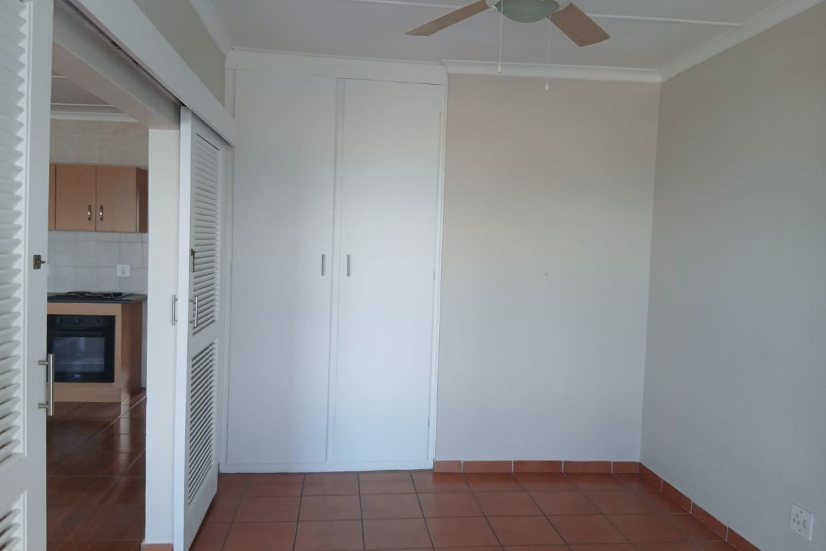 An empty bedroom featuring built-in cupboards, a ceiling fan, and tiled flooring, with a view into the adjacent kitchen area.
