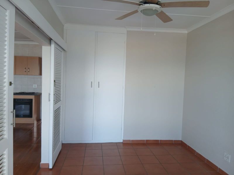 An empty bedroom featuring built-in cupboards, a ceiling fan, and tiled flooring, with a view into the adjacent kitchen area.