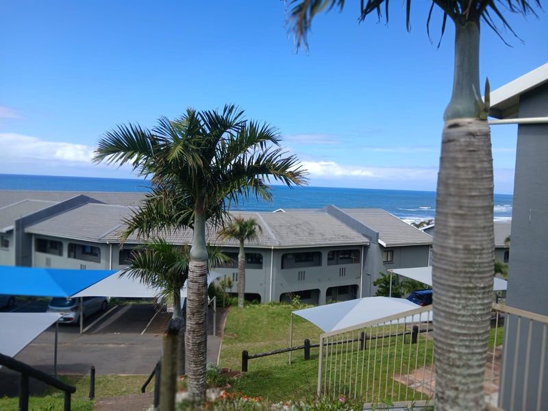 Elevated view overlooking the apartment complex grounds, featuring covered parking bays, landscaped gardens, and a scenic ocean backdrop.