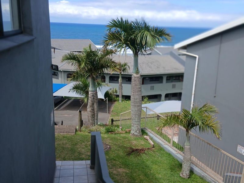 An elevated view overlooking the apartment complex grounds, featuring landscaped gardens, palm trees, and a scenic ocean backdrop.