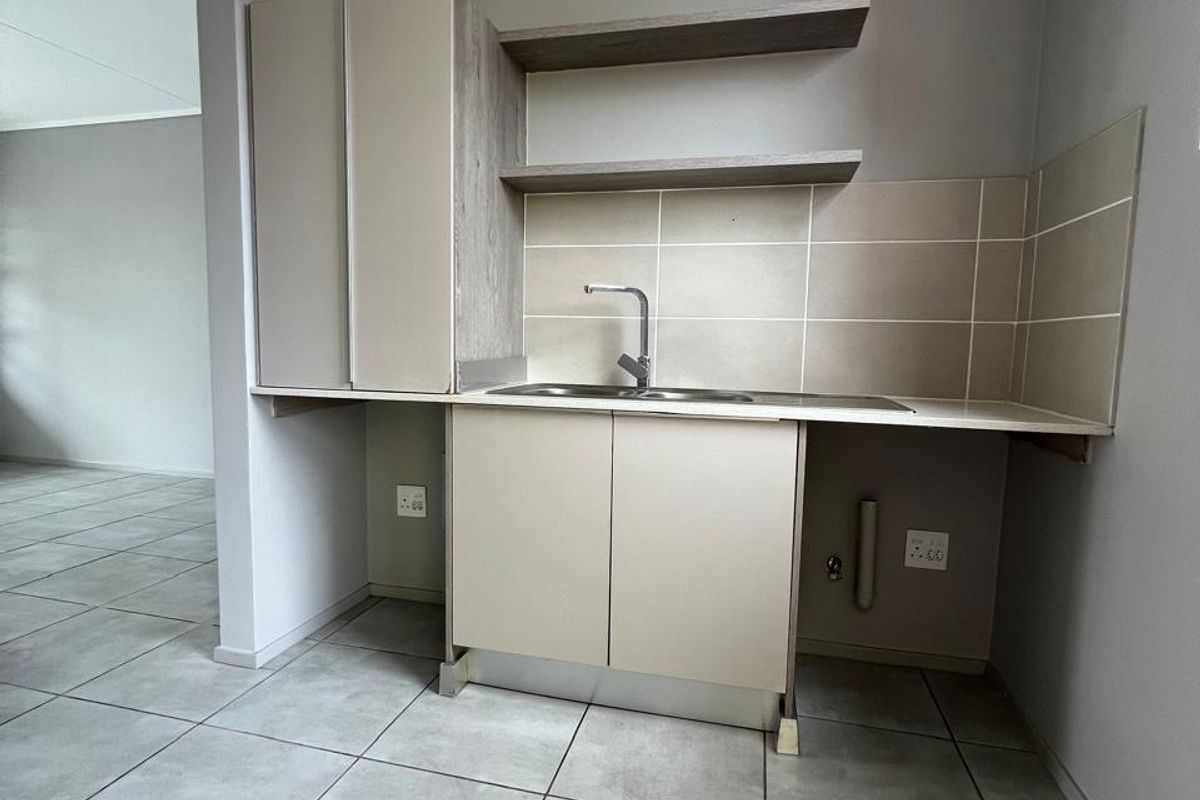 A compact, modern kitchen area featuring neutral cabinetry, open shelving, a stainless steel sink, and contemporary ceramic floor tiles in an open-plan layout.
