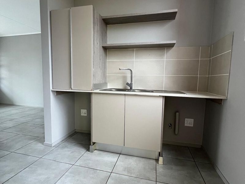 A compact, modern kitchen area featuring neutral cabinetry, open shelving, a stainless steel sink, and contemporary ceramic floor tiles in an open-plan layout.