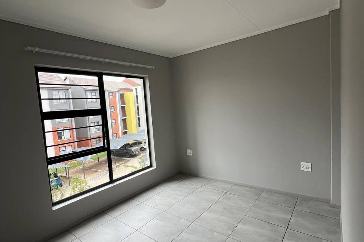 A clean, empty bedroom featuring neutral grey walls, modern tiled flooring, and a large window providing natural light and views of the complex.