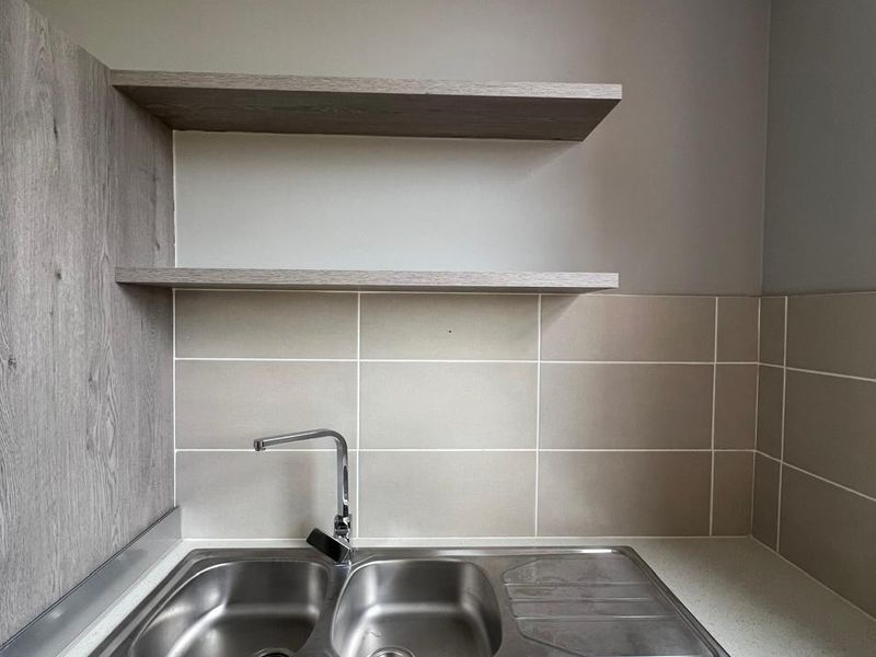 A clean and modern kitchen prep area featuring a double bowl stainless steel sink, stone-look countertop, tiled backsplash, and floating wooden shelves.