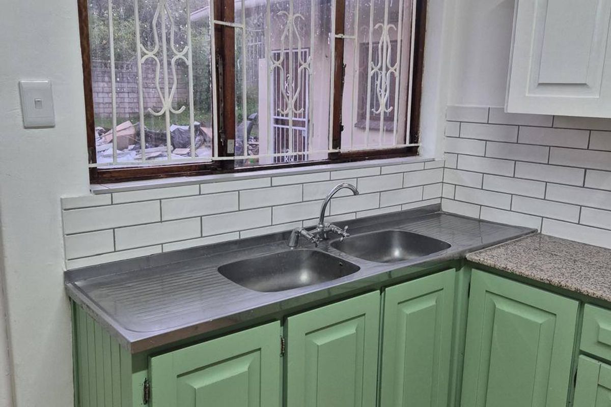 A functional kitchen featuring green cabinetry, a stainless steel double sink, subway tile backsplash, and stone countertops with a window overlooking the exterior.