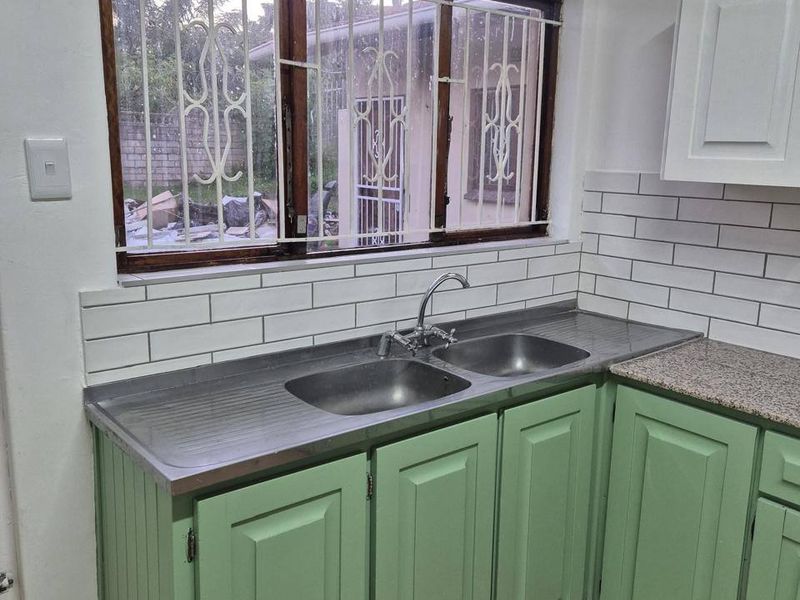A functional kitchen featuring green cabinetry, a stainless steel double sink, subway tile backsplash, and stone countertops with a window overlooking the exterior.