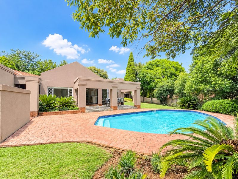 A well-maintained outdoor pool area featuring brick paving, a covered patio space, and established garden landscaping.