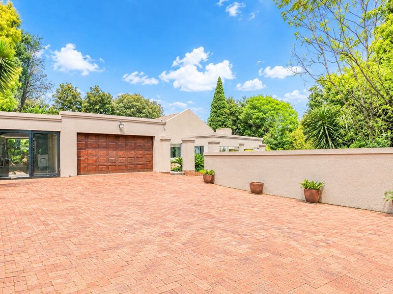 A well-maintained front exterior featuring a spacious brick-paved driveway, a secure wooden garage door, and a clean boundary wall set against a landscaped garden.