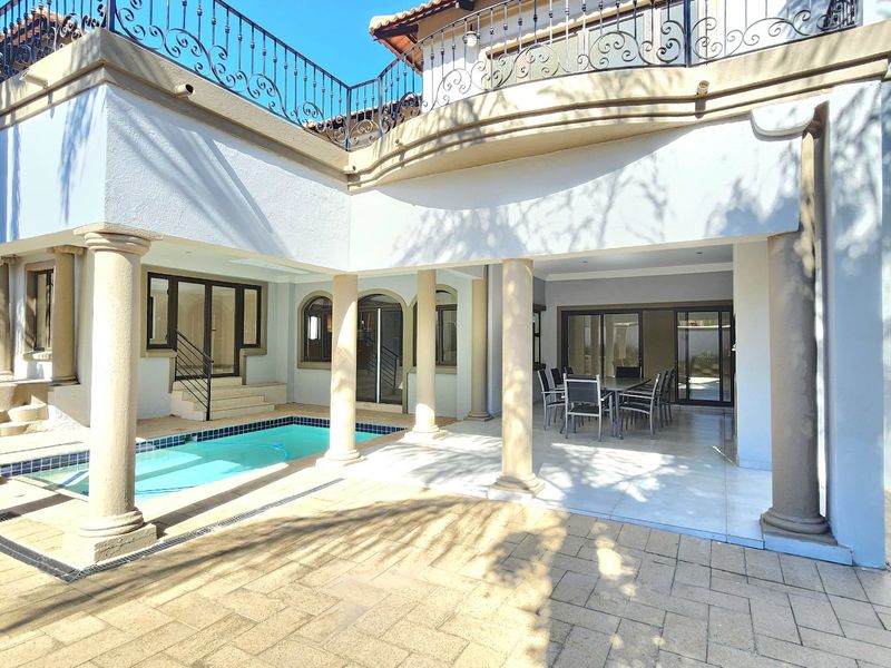 Spacious outdoor entertainment area featuring a swimming pool, covered patio with stone columns, and an upper-level balcony with decorative wrought iron railings.