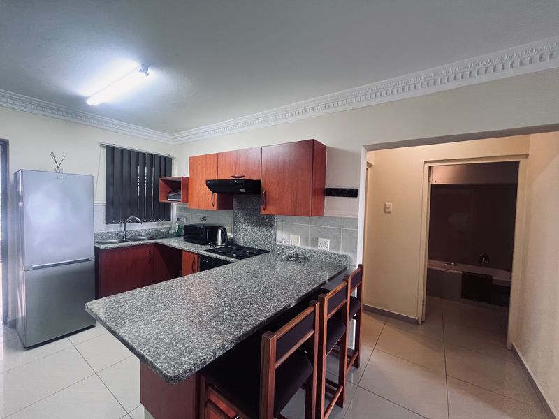 A functional kitchen featuring granite countertops, wood-finish cabinetry, a built-in hob with extractor fan, and tiled flooring, with an adjacent doorway leading to a bathroom.
