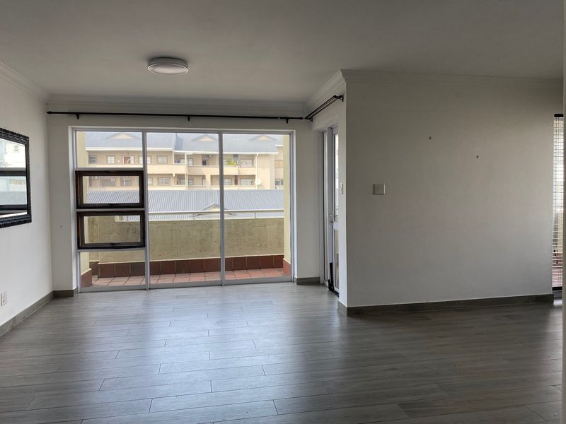 A bright, empty living room featuring modern laminate flooring, neutral walls, and large sliding glass doors providing access to an outdoor balcony area.
