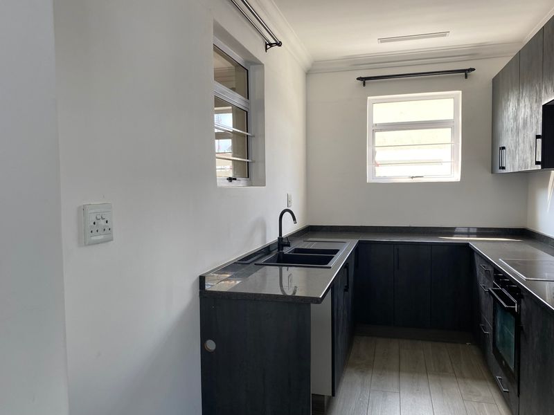 A modern, empty kitchen featuring dark cabinetry, stone countertops, a black sink with matching tap, and built-in oven and hob.