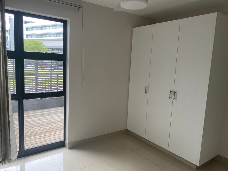 A bright, empty bedroom featuring neutral tiled flooring, a large built-in wardrobe, and a window with access to an outdoor deck area.