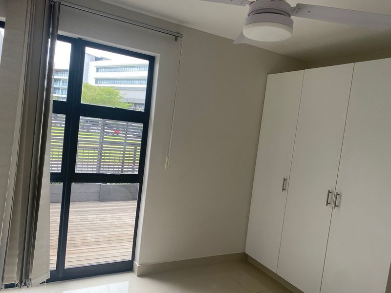 A bright, empty bedroom featuring built-in cupboards, a ceiling fan, tiled floors, and a sliding glass door leading to an outdoor deck area.