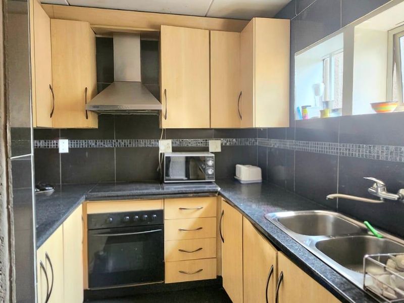A functional kitchen featuring light wood cabinetry, dark tiled backsplash, integrated oven and hob, and a double stainless steel sink.