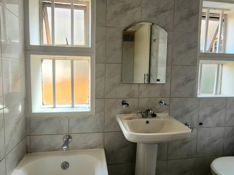 A compact bathroom featuring a pedestal basin, bathtub, and toilet, finished with marble-effect wall tiles and security bars on the windows.