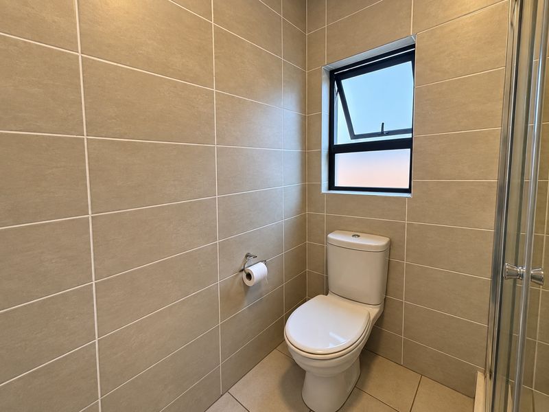 A clean, modern bathroom featuring neutral wall and floor tiles, a white toilet, and a glass-enclosed shower.