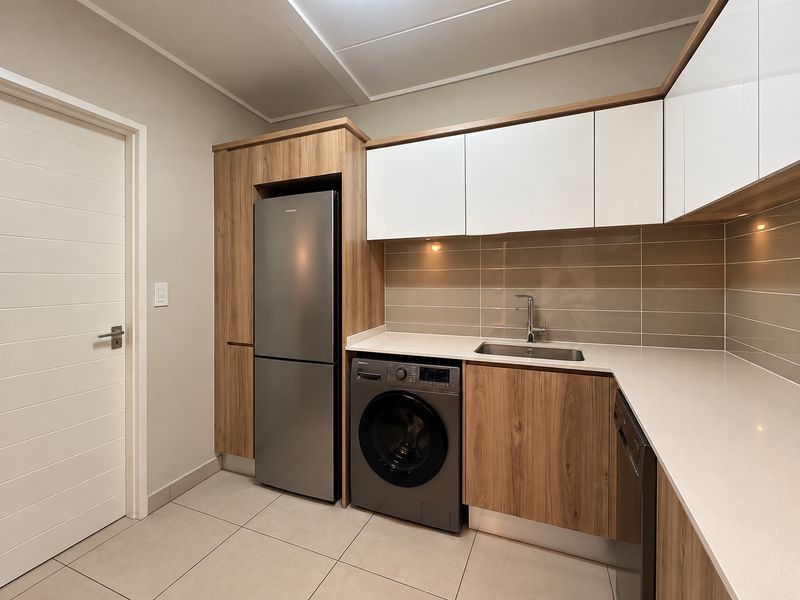 A modern, well-appointed laundry area featuring sleek wood-grain and white cabinetry, stone countertops, a stainless steel sink, and integrated appliance spaces.
