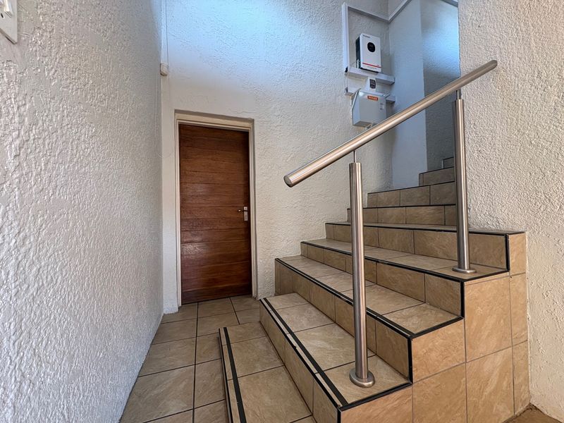 A clean hallway featuring a tiled staircase with a modern stainless steel handrail, a solid wooden door, and a wall-mounted inverter system.