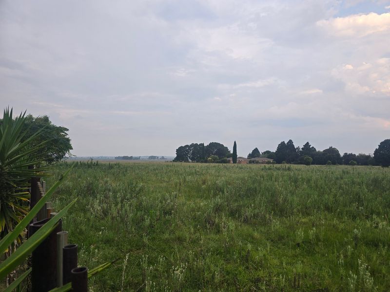 A wide-angle view of the expansive farm landscape featuring open fields and distant mature trees under a cloudy sky.