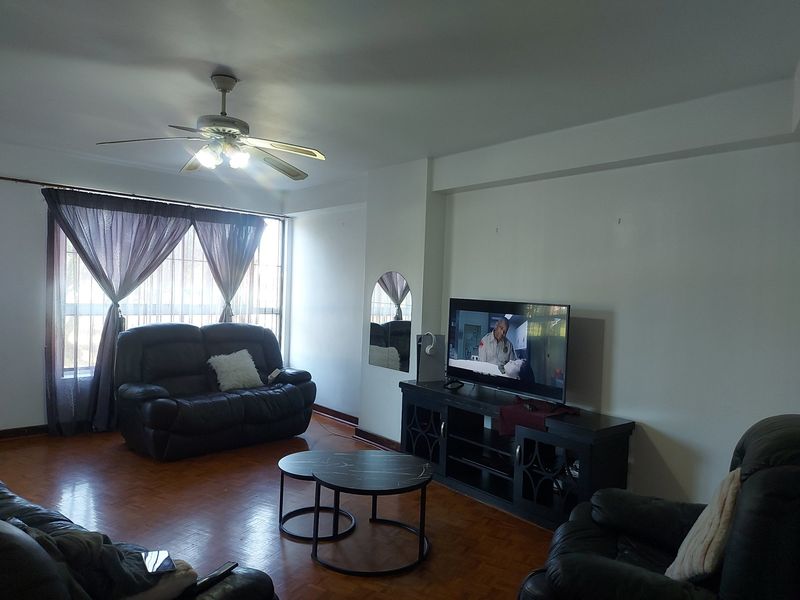 A spacious living room featuring parquet flooring, a ceiling fan, and large windows with security bars, offering a comfortable and functional living space.