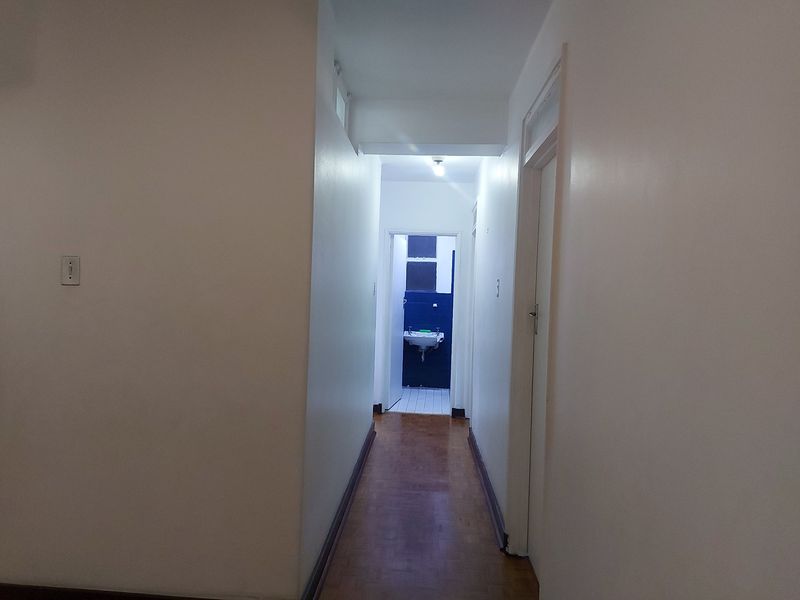 A simple, empty hallway featuring parquet flooring and access to adjacent rooms, including a view into a bathroom at the end.