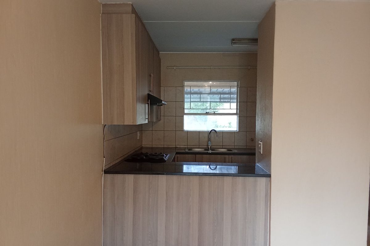 A compact, functional kitchen featuring light wood-finish cabinetry, dark countertops, a gas hob, and a tiled backsplash with a window above the sink.