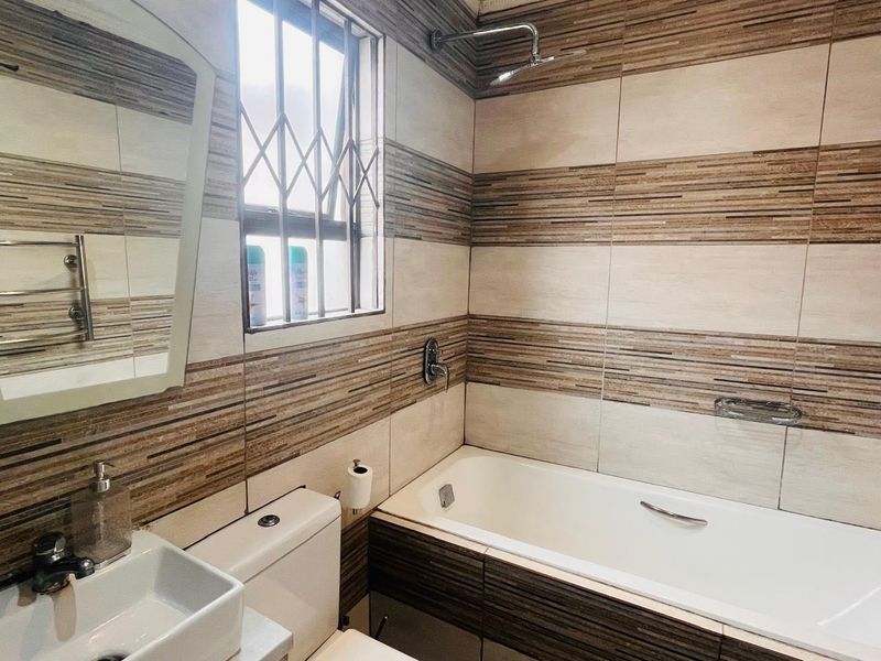 A modern bathroom featuring tiled walls, a bathtub with shower head, a wall-mounted basin, and a toilet with security bars on the window.
