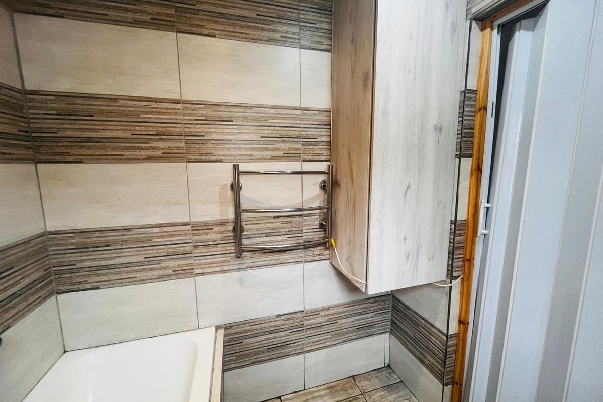 A bathroom featuring tiled walls and flooring, a built-in wall cabinet, and a heated towel rail, with a bathtub visible in the corner.