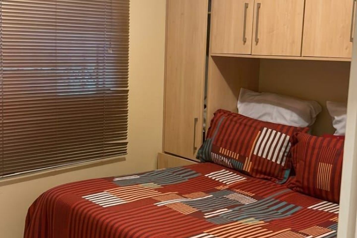 A compact bedroom featuring built-in wooden cupboards, tiled flooring, and venetian blinds, providing functional storage and natural light control.