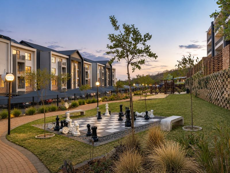 A well-maintained communal garden area featuring a large outdoor chess set, paved pathways, and landscaped grounds within a modern apartment complex.