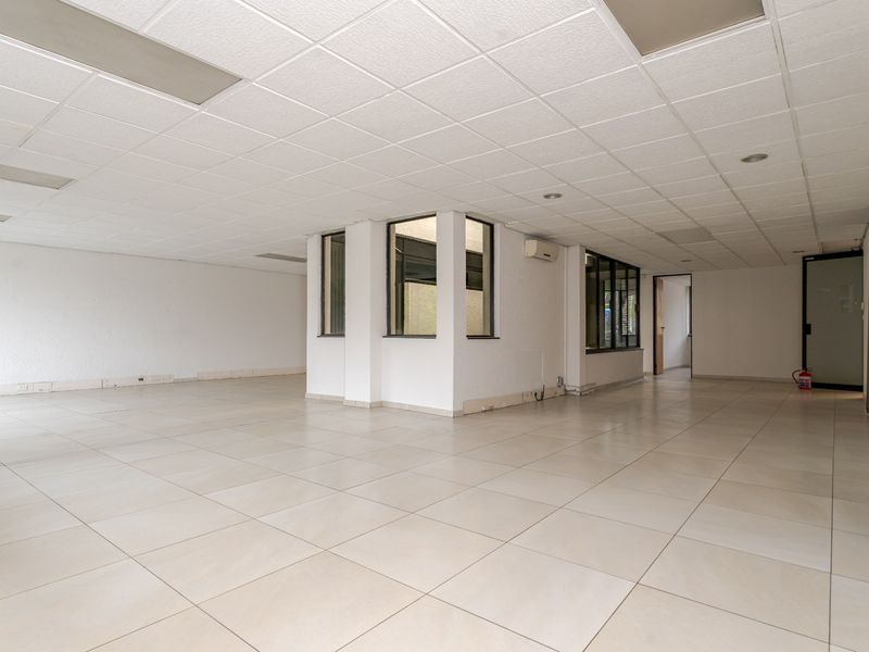 A spacious, empty open-plan commercial office area featuring tiled flooring, a suspended ceiling with recessed lighting, and integrated air conditioning.