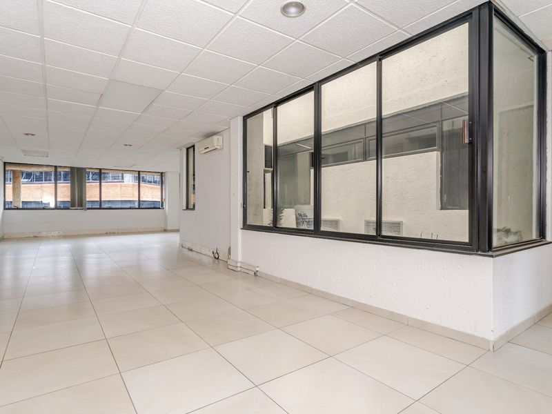 An empty, open-plan commercial office space featuring tiled flooring, a suspended ceiling, and a large glass partition wall.