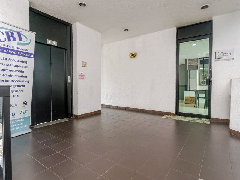A commercial building entrance lobby featuring tiled flooring, a modern elevator, and a glass-paneled door leading into an office space.