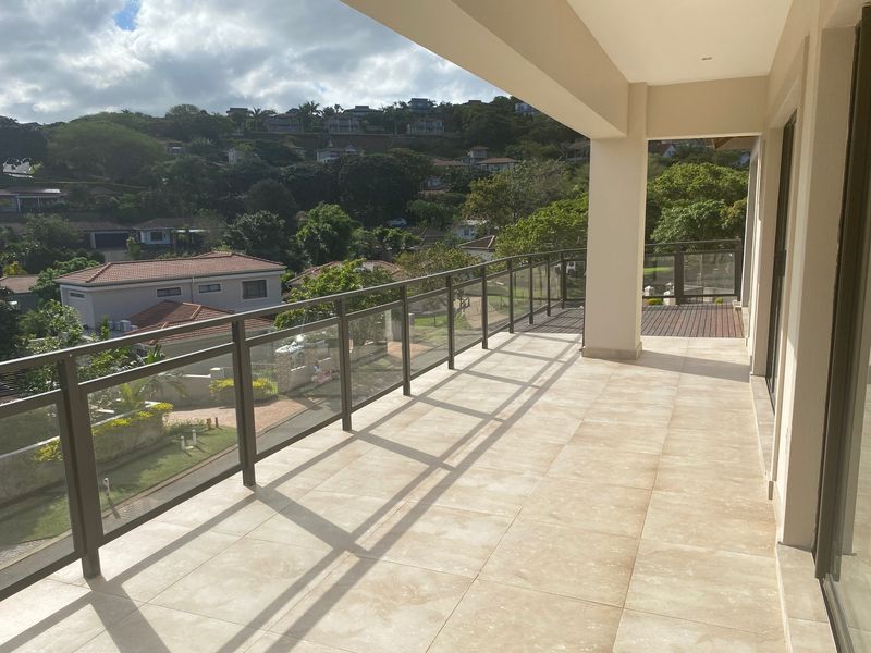 A spacious, covered balcony featuring modern tiled flooring and a sleek glass balustrade, offering elevated views of the surrounding residential neighborhood.
