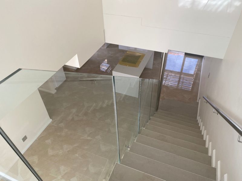 A modern staircase featuring a sleek glass balustrade, stainless steel handrail, and neutral tiled flooring, leading down to an open-plan living area.