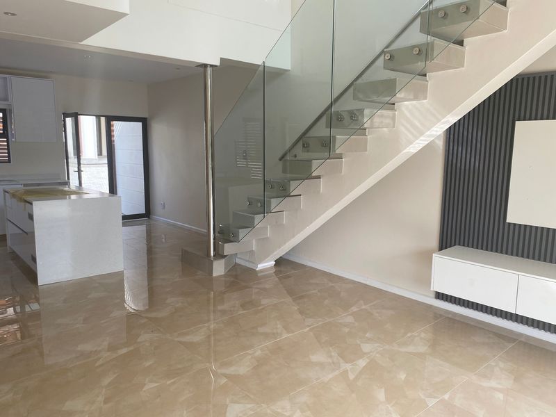 Modern open-plan living area featuring a striking floating staircase with glass balustrade, sleek kitchen island, and contemporary tiled flooring throughout.