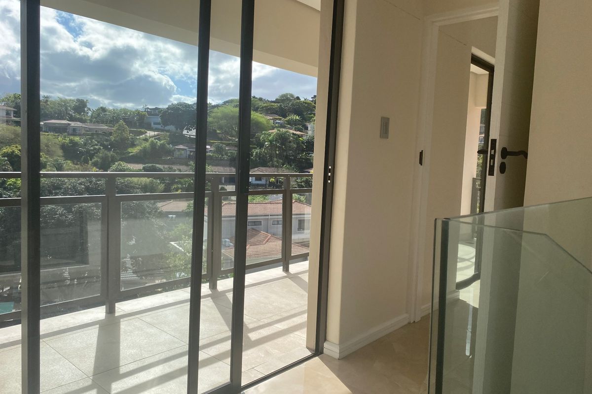 A bright balcony area featuring tiled flooring, a modern glass balustrade, and sliding glass doors leading to an interior hallway.
