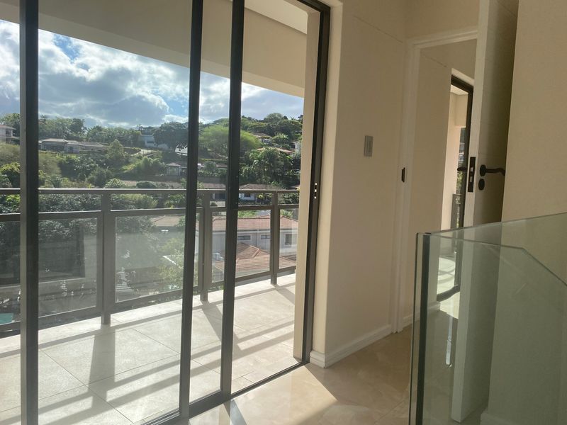 A bright balcony area featuring tiled flooring, a modern glass balustrade, and sliding glass doors leading to an interior hallway.