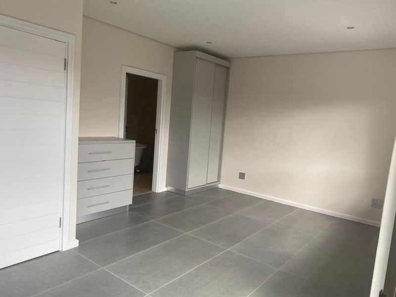A clean, modern bedroom featuring large format grey floor tiles, recessed lighting, and a built-in wardrobe with direct access to an en-suite bathroom.