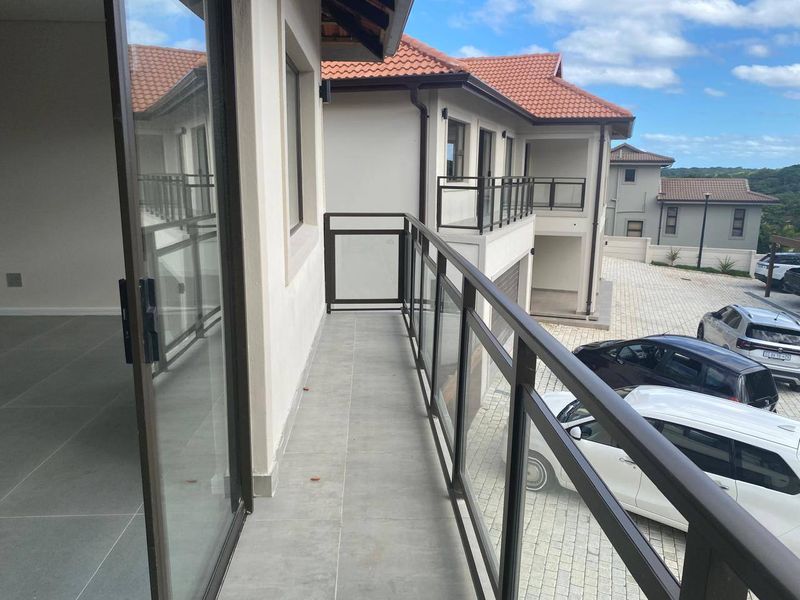 A modern balcony featuring a glass balustrade and tiled flooring, providing an elevated outdoor space overlooking the apartment complex parking area.
